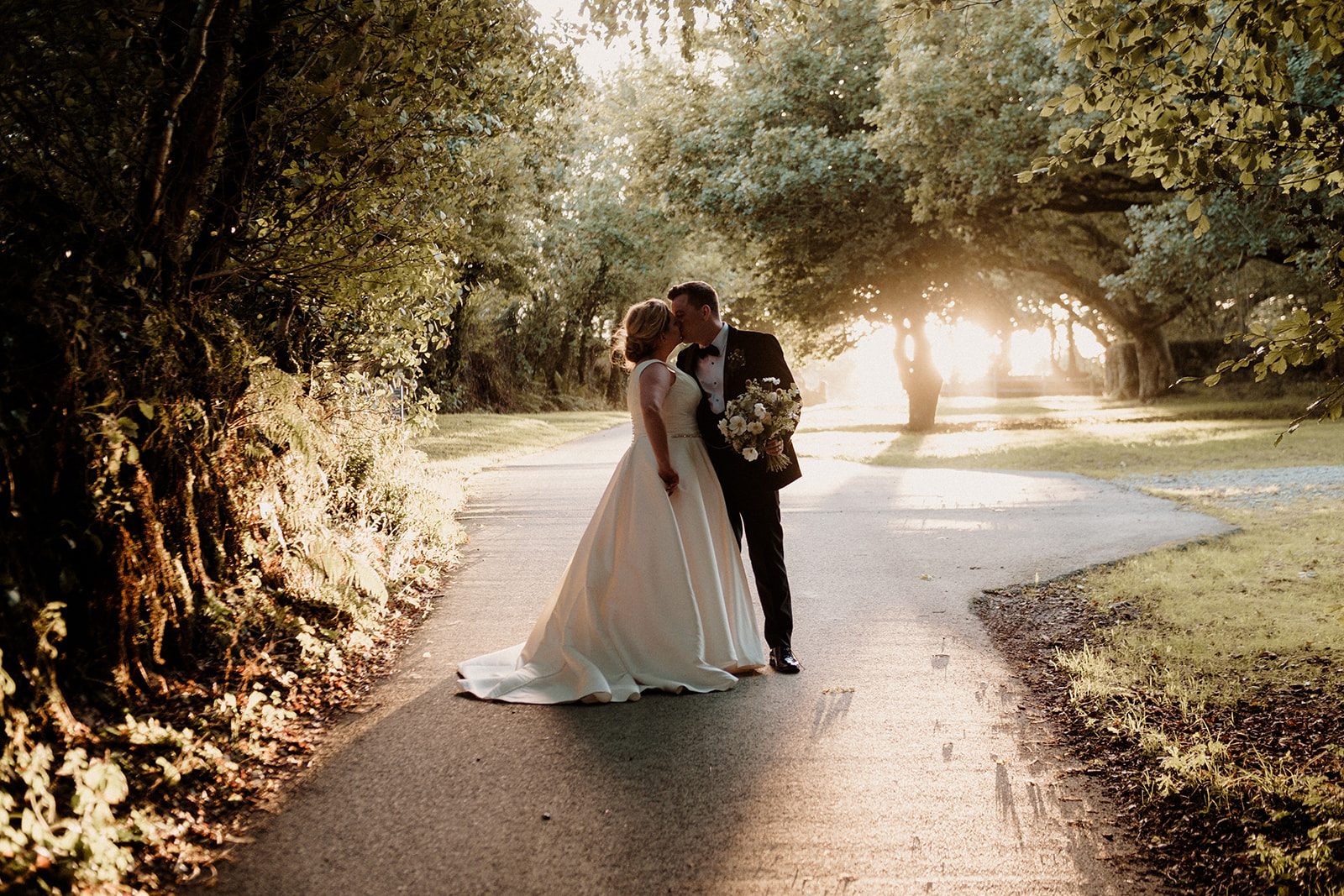 Amy and David kissing on a tree-lined lane in golden evening light