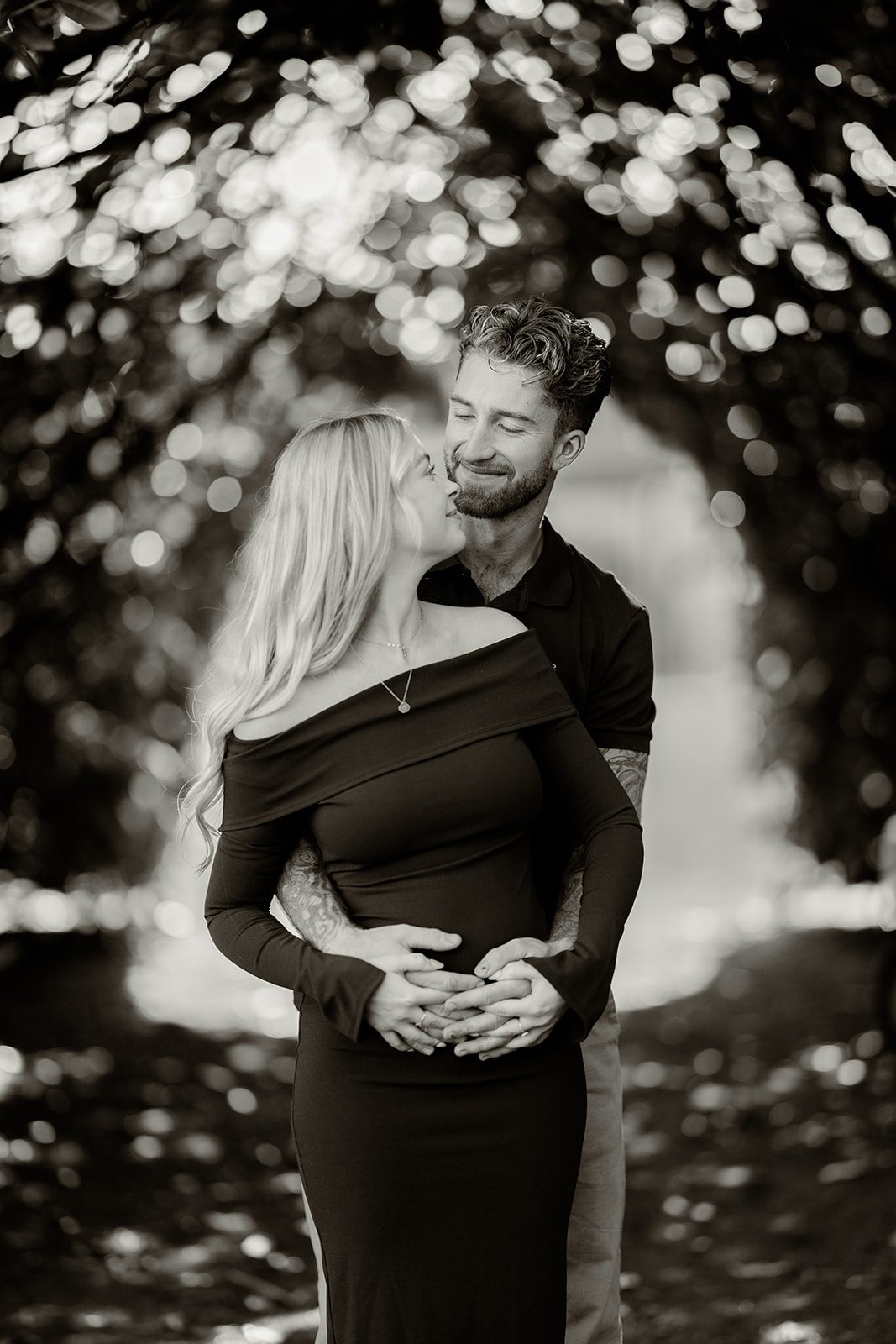 A couple embracing beneath trees during their maternity shoot, in black and white