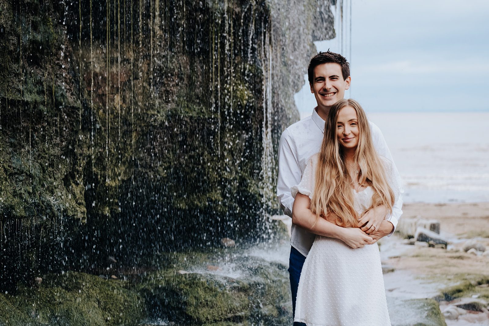 An engaged couple embracing beside the waterfall at St Audries Bay