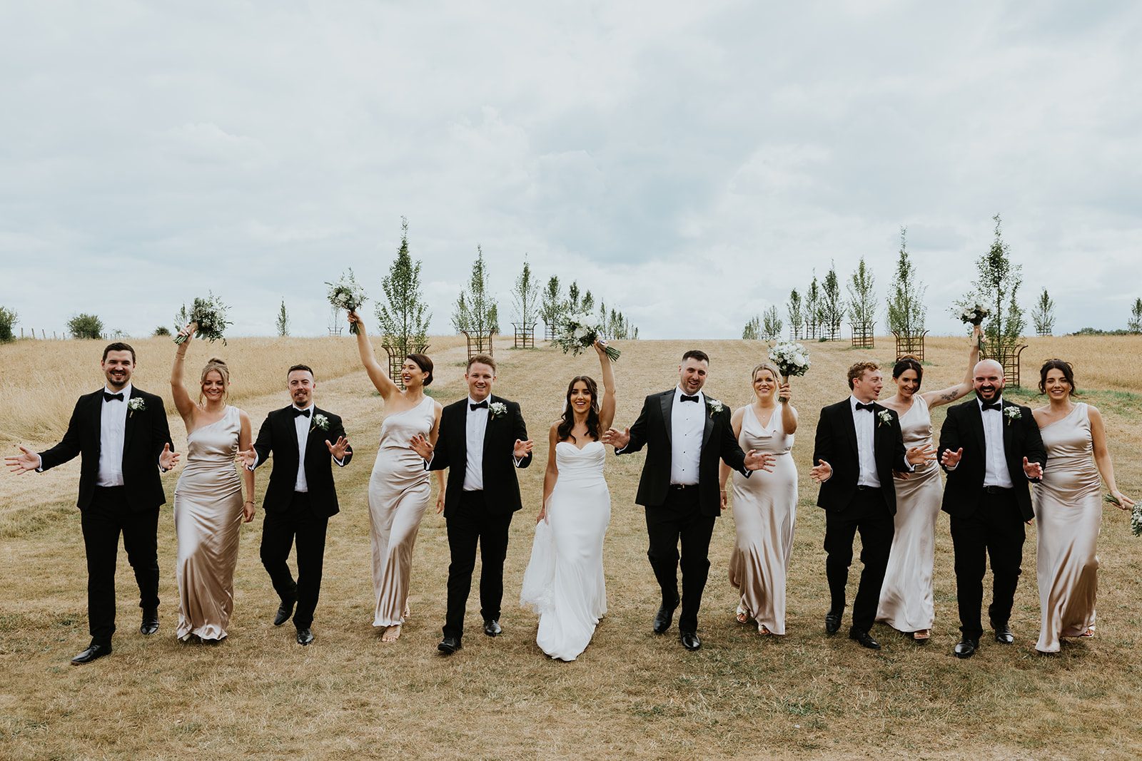 Gemma and Matt celebrating in a field with their wedding party and bouquets raised