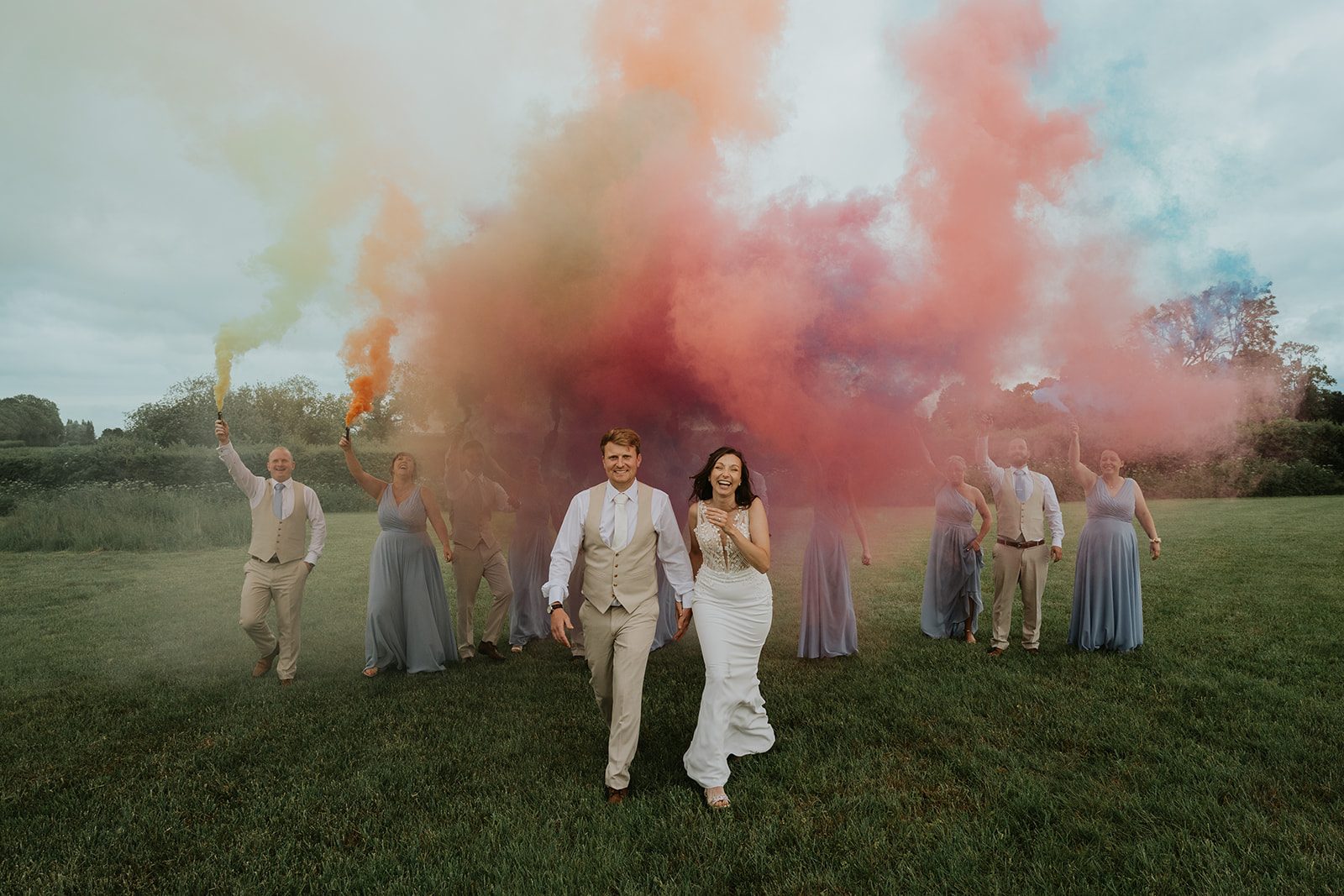 Jade and Paul laughing with their wedding party beneath clouds of colourful smoke