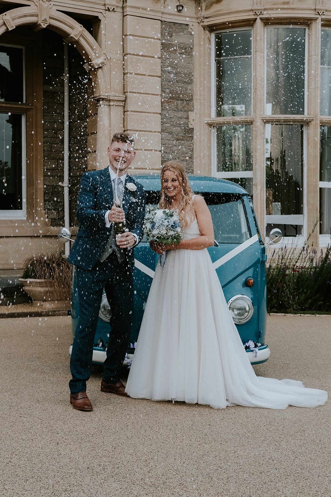 Jessie and Kyle opening champagne beside a blue wedding camper van