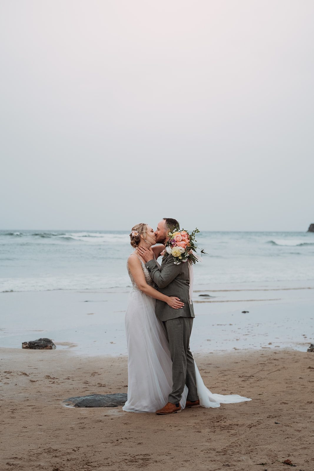 Kirsty and Kester kissing on the beach with a colourful bouquet