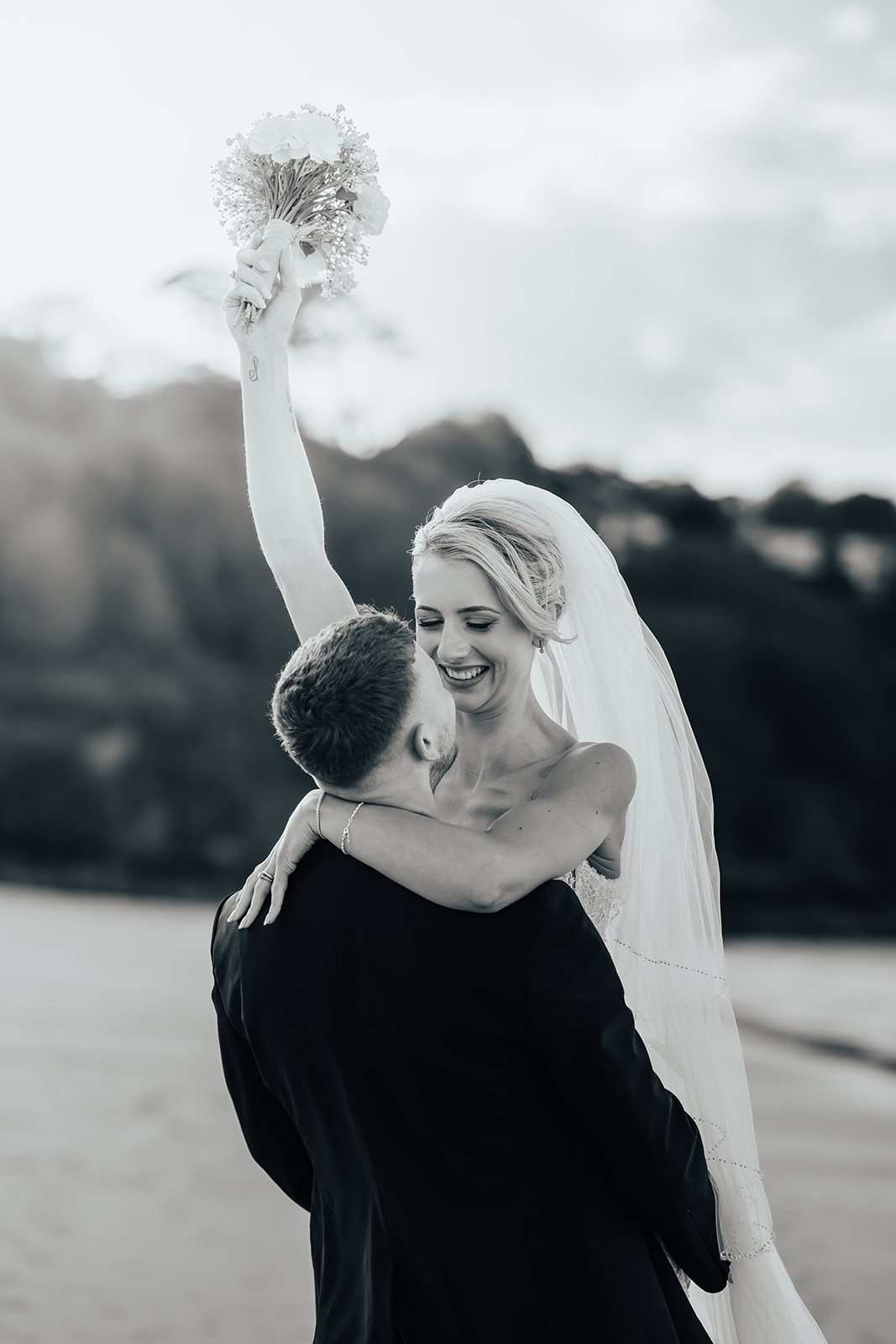 Maisie laughing as Pete lifts her, with her bouquet in the air, in black and white
