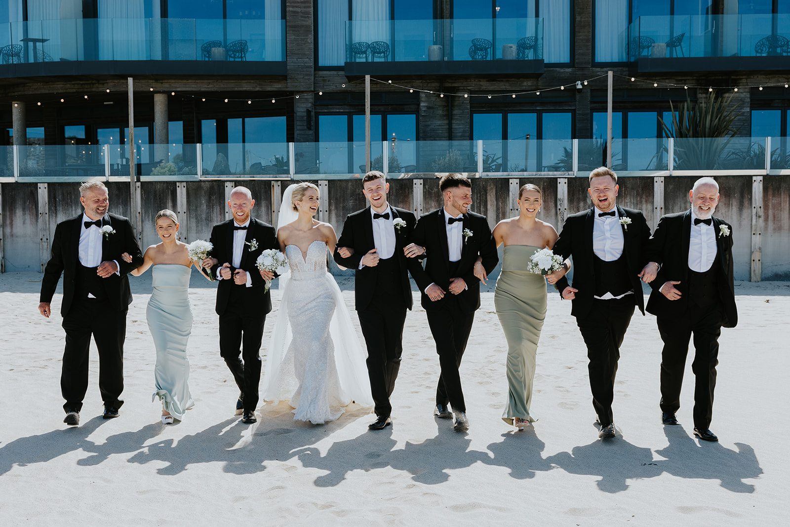 Maisie and Pete laughing as their bridal party walks together on the beach