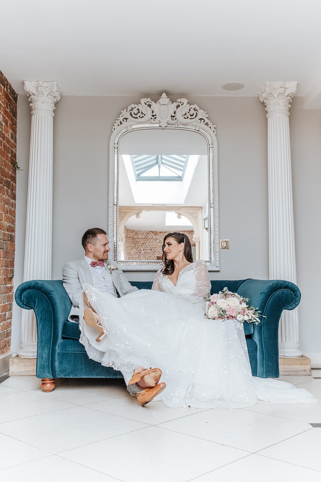 Sophie and Callum smiling at each other on a teal sofa beneath an ornate mirror